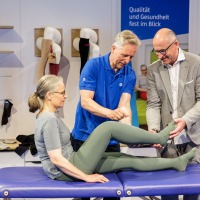 Eine Frau liegt auf einer Behandlungsliege auf der OTWorld, während zwei Männer – einer im blauen Poloshirt, der andere im grauen Anzug – ihre Beine in grüner Kompressionskleidung für eine Demonstration halten; im Hintergrund sind Strümpfe an Wandhalterungen sowie ein Schild mit der Aufschrift „Qualität und Gesundheit fest im Blick“ zu sehen. Eine Frau liegt auf einer Behandlungsliege auf der OTWorld, während zwei Männer – einer im blauen Poloshirt, der andere im grauen Anzug – ihre Beine in grüner Kompressionskleidung für eine Demonstration halten; im Hintergrund sind Strümpfe an Wandhalterungen sowie ein Schild mit der Aufschrift „Qualität und Gesundheit fest im Blick“ zu sehen.