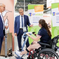 Eine junge Frau sitzt im Rollstuhl und nutzt auf der OTWorld ein Therapiegerät mit Pedalen, während zwei Männer in Anzügen das Gerät begutachten; im Hintergrund stehen zwei grüne OTWorld-Roll-Up-Banner mit englischer und deutscher Aufschrift. Eine junge Frau sitzt im Rollstuhl und nutzt auf der OTWorld ein Therapiegerät mit Pedalen, während zwei Männer in Anzügen das Gerät begutachten; im Hintergrund stehen zwei grüne OTWorld-Roll-Up-Banner mit englischer und deutscher Aufschrift.