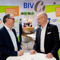 Zwei männliche Personen unterhalten sich an einem Stehtisch. Im Hintergrund ist das Logo der OTWorld auf einem Aufsteller zu sehen. Zwei männliche Personen unterhalten sich an einem Stehtisch. Im Hintergrund ist das Logo der OTWorld auf einem Aufsteller zu sehen.