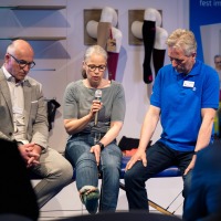 Eine Frau mit Mikrofon sitzt zwischen zwei Männern und redet über Kompressionsstrümpfe. Eine Frau mit Mikrofon sitzt zwischen zwei Männern und redet über Kompressionsstrümpfe.