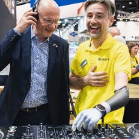 Mann mit Prothesenarm erklärt DJ-Equipment, während ein anderer Mann mit Kopfhörern zuhört und lächelt. Mann mit Prothesenarm erklärt DJ-Equipment, während ein anderer Mann mit Kopfhörern zuhört und lächelt.