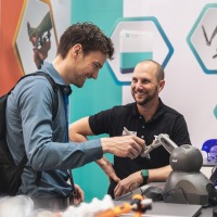 Messebesucher testet ein technisches Assistenzsystem am Ausstellungsstand, während ein Aussteller ihm lächelnd zusieht. Messebesucher testet ein technisches Assistenzsystem am Ausstellungsstand, während ein Aussteller ihm lächelnd zusieht.