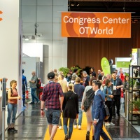 Menschen gehen durch eine Messehalle mit orangefarbenem Teppich. Im Hintergrund hängt ein großes oranges Banner mit der Aufschrift „Congress Center OTWorld“. Links ist ein großer Werbeaufsteller mit dem Bild einer Frau zu sehen. Besucherinnen und Besucher bewegen sich zwischen den Ständen und unterhalten sich. Menschen gehen durch eine Messehalle mit orangefarbenem Teppich. Im Hintergrund hängt ein großes oranges Banner mit der Aufschrift „Congress Center OTWorld“. Links ist ein großer Werbeaufsteller mit dem Bild einer Frau zu sehen. Besucherinnen und Besucher bewegen sich zwischen den Ständen und unterhalten sich.