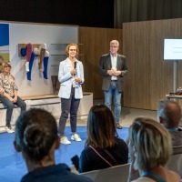 Eine Frau spricht auf einer kleinen Bühne vor einem sitzenden Publikum in ein Mikrofon. Ein Mann steht neben ihr, während zwei weitere Frauen im Hintergrund sitzen. Medizinische Kompressionsstrümpfe sind an den Beinen von Schaufensterpuppen ausgestellt, die an der Wand befestigt sind. Auf der rechten Seite sind ein Bildschirm und ein Tisch mit Ausstellungsstücken zu sehen. Eine Frau spricht auf einer kleinen Bühne vor einem sitzenden Publikum in ein Mikrofon. Ein Mann steht neben ihr, während zwei weitere Frauen im Hintergrund sitzen. Medizinische Kompressionsstrümpfe sind an den Beinen von Schaufensterpuppen ausgestellt, die an der Wand befestigt sind. Auf der rechten Seite sind ein Bildschirm und ein Tisch mit Ausstellungsstücken zu sehen.