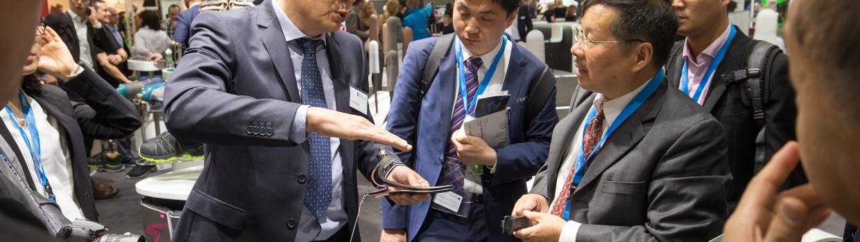 A man in a suit explains a technical product to a group of trade fair visitors at an exhibition booth. A man in a suit explains a technical product to a group of trade fair visitors at an exhibition booth.